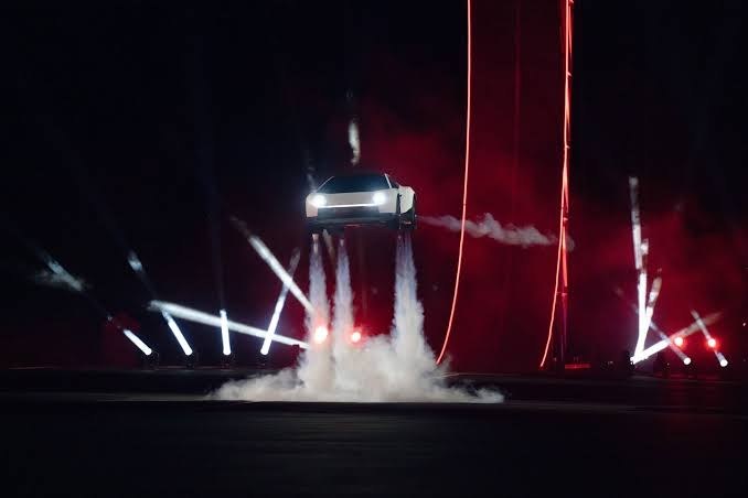 El Roadster blanco suspendido en el aire sobre cuatro columnas de humo de sus propulsores, en un escenario rojo con focos.